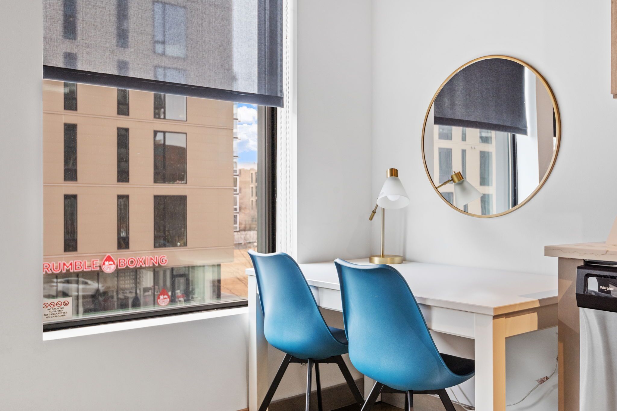 A room with a white desk, two blue chairs, a round mirror, and a window with a view of buildings and a sign reading "RUMBLE BOXING".