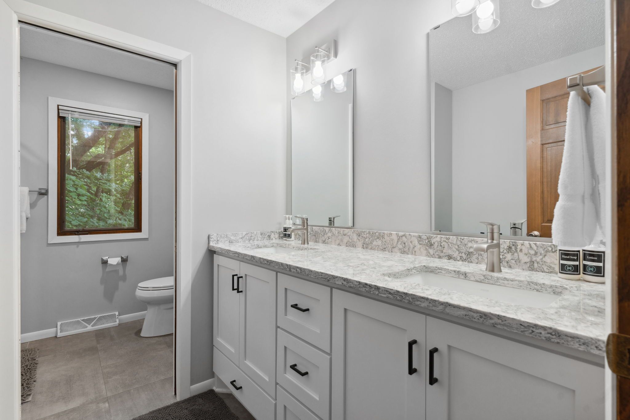 A bathroom with a white vanity, a large mirror, and a window with a view of trees.