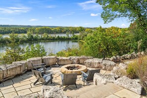 An outdoor seating area with stone tables and chairs, a fire pit, and a view of a lake surrounded by trees.