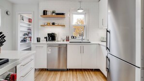 A modern kitchen with white cabinetry, a stainless steel dishwasher, and a built-in oven. There are shelves above the sink holding various items, and a window above the sink provides natural light. - Nokomis Blue (Minneapolis)