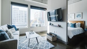 A modern hotel room with a large window, a flat-screen TV, a bed, a sofa, and a coffee table. - Sable 206 - Alcove (Minneapolis)