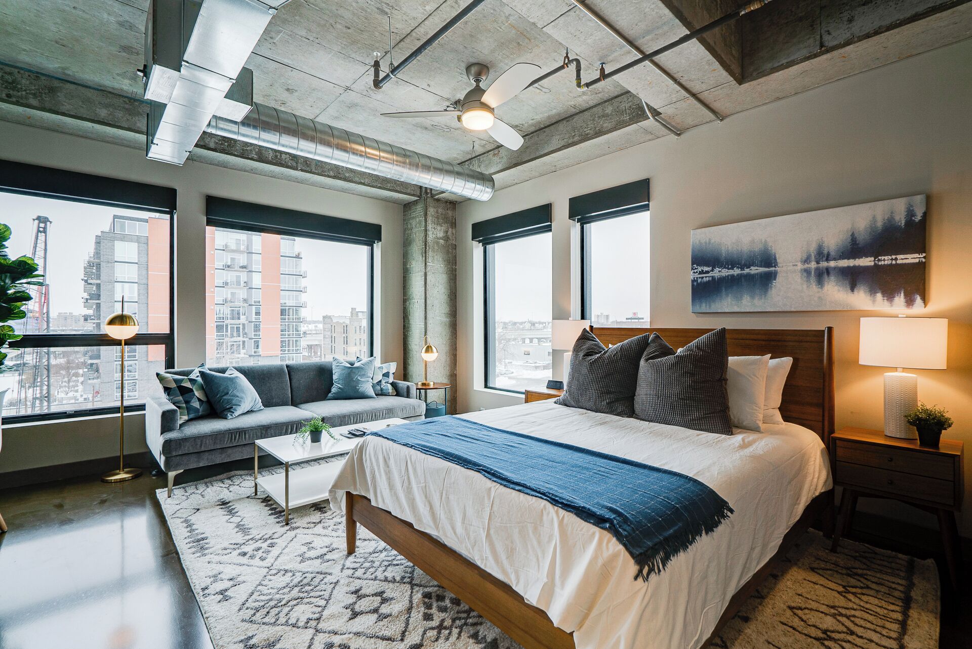 A modern hotel room with a large bed, a sofa, and a view of the city through large windows.