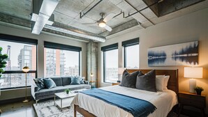 A modern hotel room with a large bed, a sofa, and a view of the city through large windows. - Sable 401 - Studio (Minneapolis)