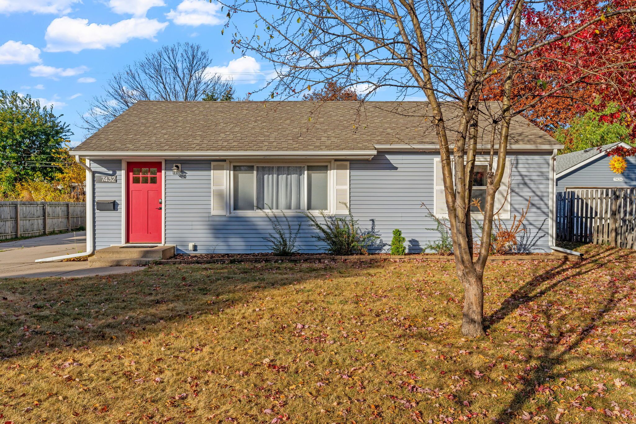 A single-story house with a red door, a number '7432' on the front, and a small front yard with grass and a tree.