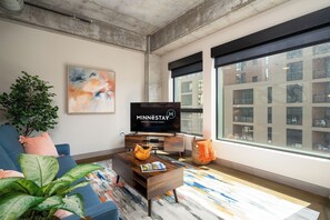A modern living room with a large window, a flat-screen TV, a sofa, a coffee table, and a potted plant. - Sable 305 - One Bedroom (Minneapolis)