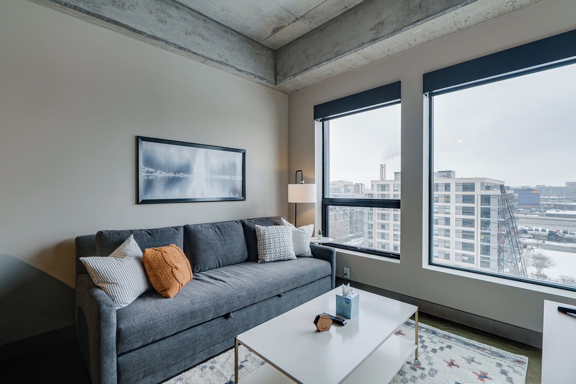 Ein modernes Wohnzimmer mit einem grauen Sofa, einem gerahmten Bild und einem großen Fenster mit Blick auf die Stadt.