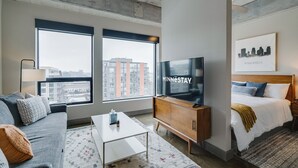 A modern hotel room with a large TV, a sofa, a bed, and a view of the city through large windows. - Sable 703 - Alcove (Minneapolis)
