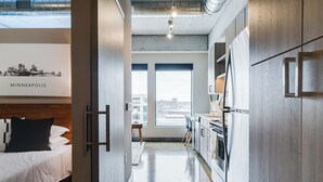 Een moderne hotelkamer met een bed, kitchenette en grote ramen met uitzicht op de stad.