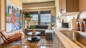 Ein modernes Wohnzimmer mit einer Couch, einem Couchtisch und einer Bank. An der Wand hängt ein Gemälde, eine Pflanze steht bereit und es gibt einen Blick auf die Stadt durch große Fenster.