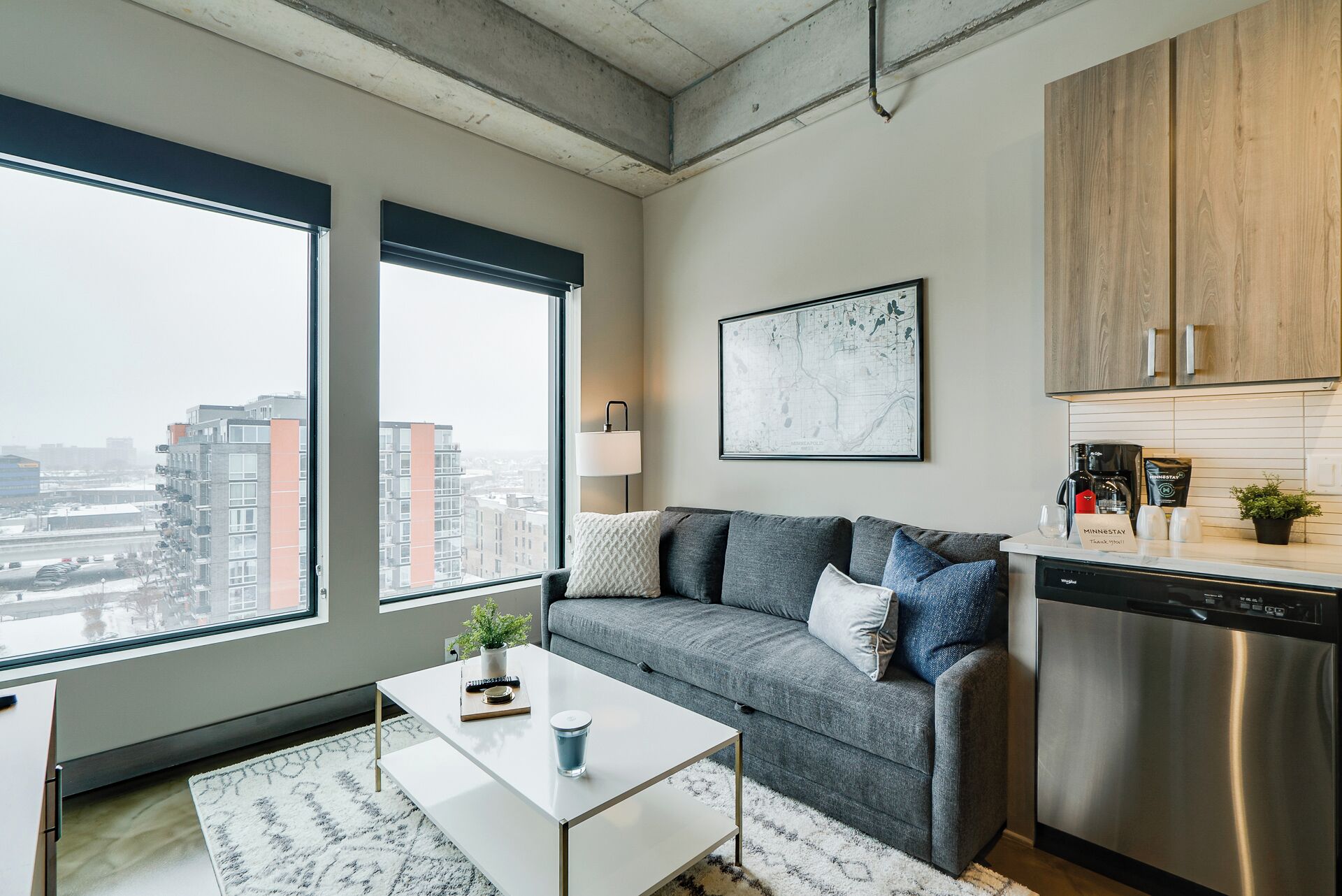 Un appartement moderne avec un canapé gris, une table basse blanche et une grande fenêtre donnant sur la ville.