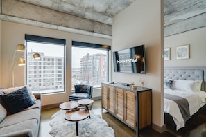 A modern hotel room with a large window, a flat-screen TV, a sofa, a chair, a small coffee table, a cabinet, and a bed. - Sable 507 - Alcove (Minneapolis)