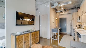 A modern hotel room with a large flat-screen TV, wooden cabinets, a kitchenette, and a dining area.
