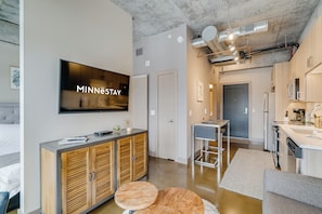 A modern hotel room with a large flat-screen TV, wooden cabinets, a kitchenette, and a dining area. - Sable 507 - Alcove (Minneapolis)