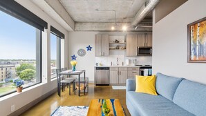 A modern living room with a sofa, coffee table, and dining area. A kitchen is visible in the background with cabinets and appliances. - Sable 604 - One Bedroom (Minneapolis)