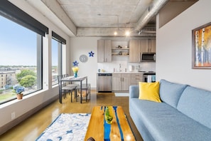 A modern living room with a sofa, coffee table, and dining area. A kitchen is visible in the background with cabinets and appliances. - Sable 604 - One Bedroom (Minneapolis)
