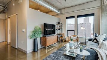 A modern living room with a large flat-screen TV, a sofa, a coffee table, and a large window with a city view.