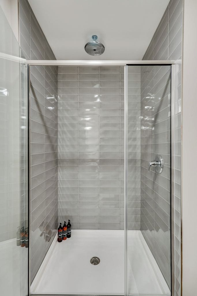Ducha con mampara de vidrio, azulejos grises, un cabezal de ducha y tres botellas de champú en el suelo.