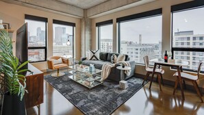 A modern living room with a sofa, coffee table, and chairs, large windows with a city view, and a plant. - Sable 608 - One Bedroom (Minneapolis)