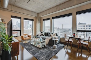 A modern living room with a sofa, coffee table, and chairs, large windows with a city view, and a plant. - Sable 608 - One Bedroom (Minneapolis)