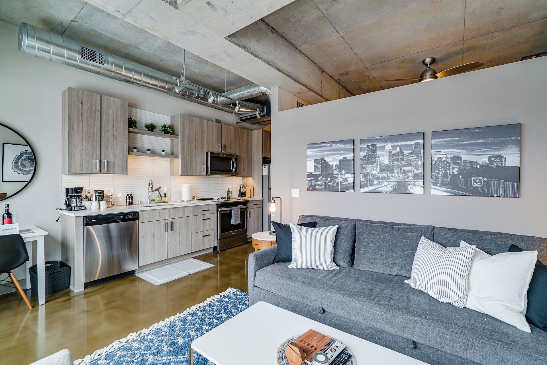 Een moderne woonruimte met een keuken, een grijze bank met gestreepte kussens en stadsgezicht kunst aan de muur.