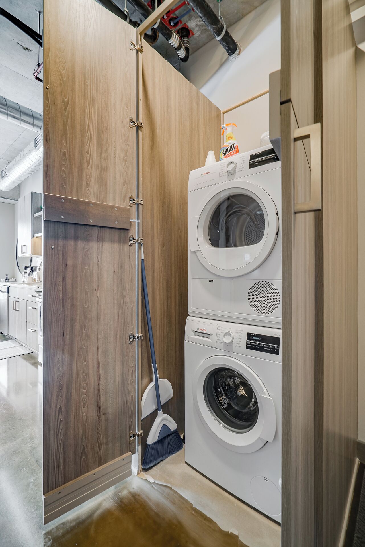 A compact laundry room with a Bosch washing machine and dryer, a wooden door, and a broom leaning against the wall.