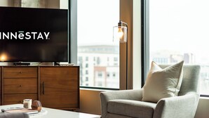 A modern hotel room with a large flat-screen TV, a comfortable armchair, a coffee table, and a view of the city through large windows. - Sable 606 - One Bedroom (Minneapolis)