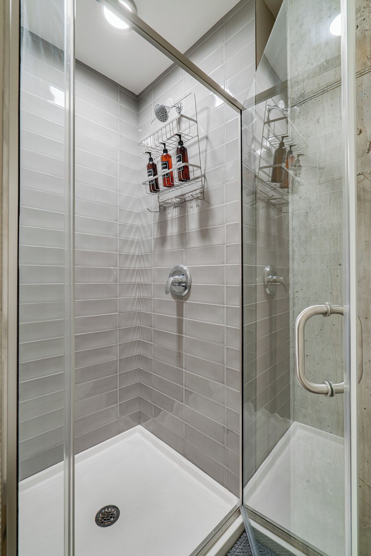A glass-enclosed shower with a white tiled wall, a showerhead, and a handrail.