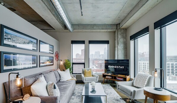 A modern living room with a large TV, comfortable seating, framed cityscape photos, and large windows offering a city view. - Sable 807 - One Bedroom (Minneapolis)