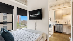 Egy modern apartman, melyben egy nagy ágy, a falra szerelt televízió, fa szekrényekkel berendezett konyha található, és kilátás nyílik az ablakon a környező épületekre.