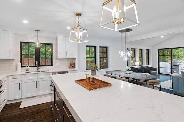 A modern kitchen with white cabinets, a large island, pendant lights, and a dining area with a table and chairs.