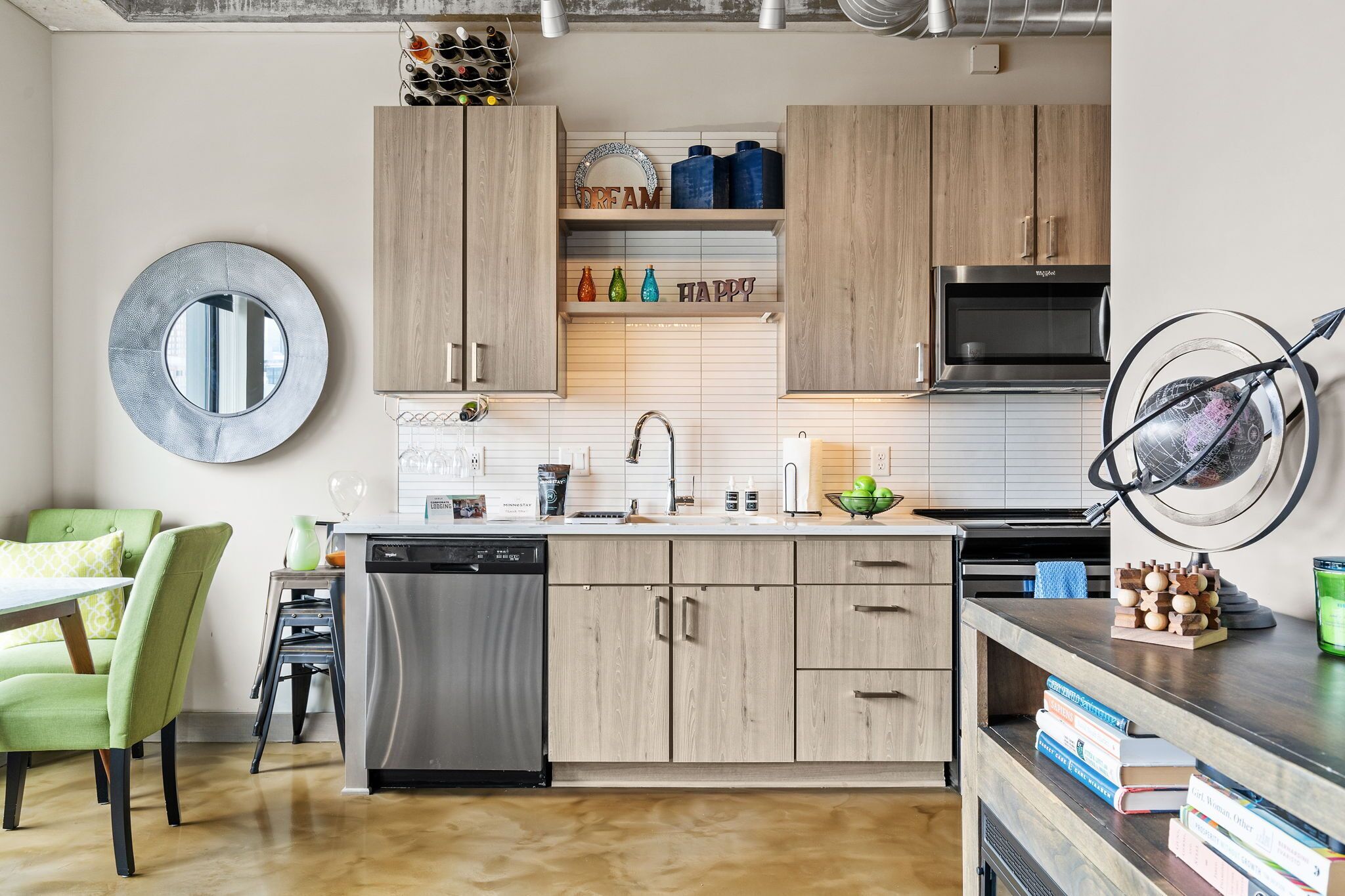Una cocina moderna con armarios de madera, un lavavajillas de acero inoxidable y un microondas. Hay una silla verde y una mesita con una lámpara.