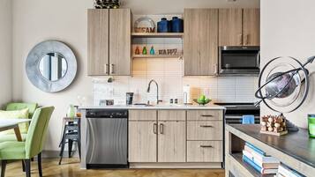 Una cocina moderna con armarios de madera, un lavavajillas de acero inoxidable y un microondas. Hay una silla verde y una mesita con una lámpara.
