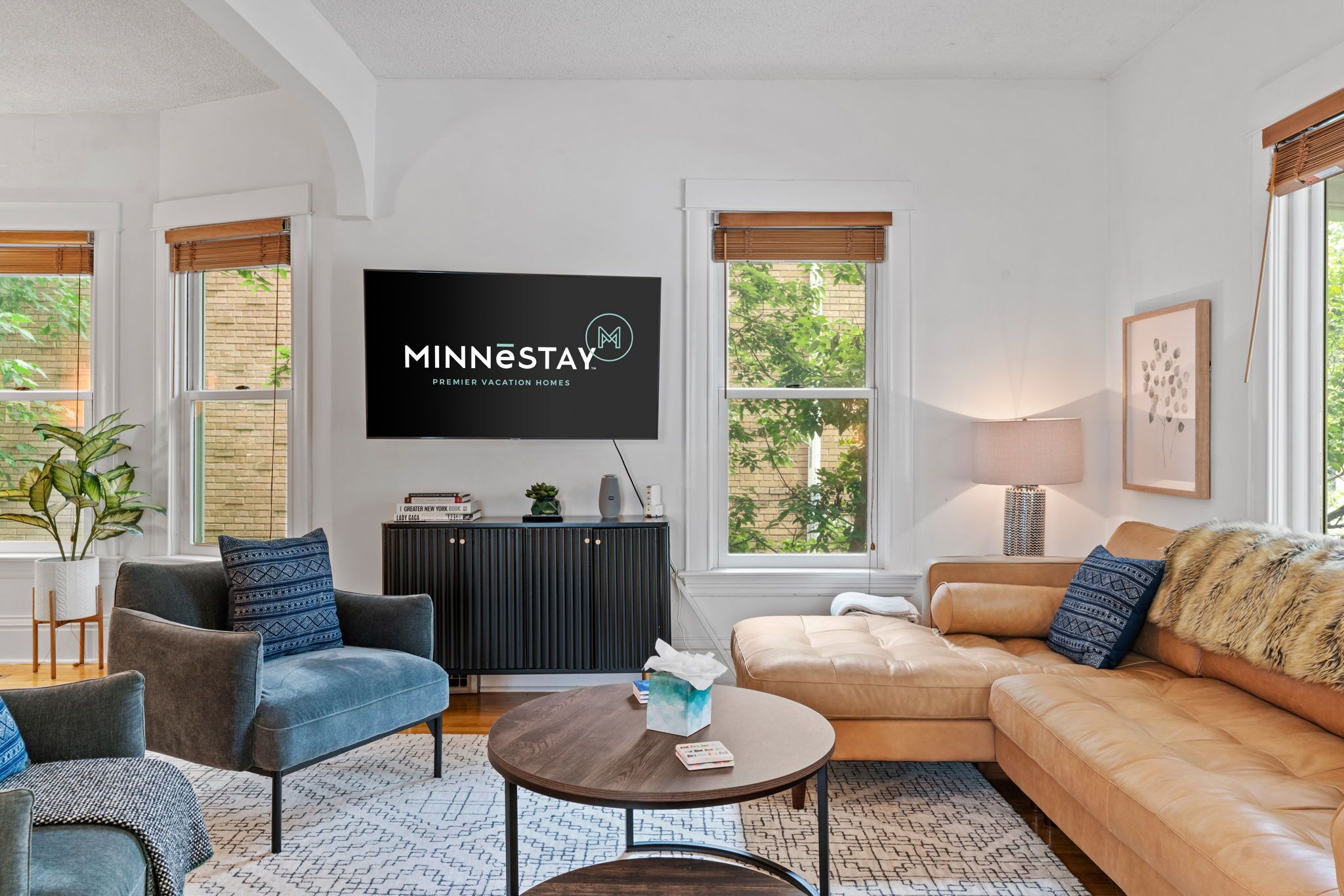 A modern living room with a flat-screen TV, comfortable seating, and large windows.
