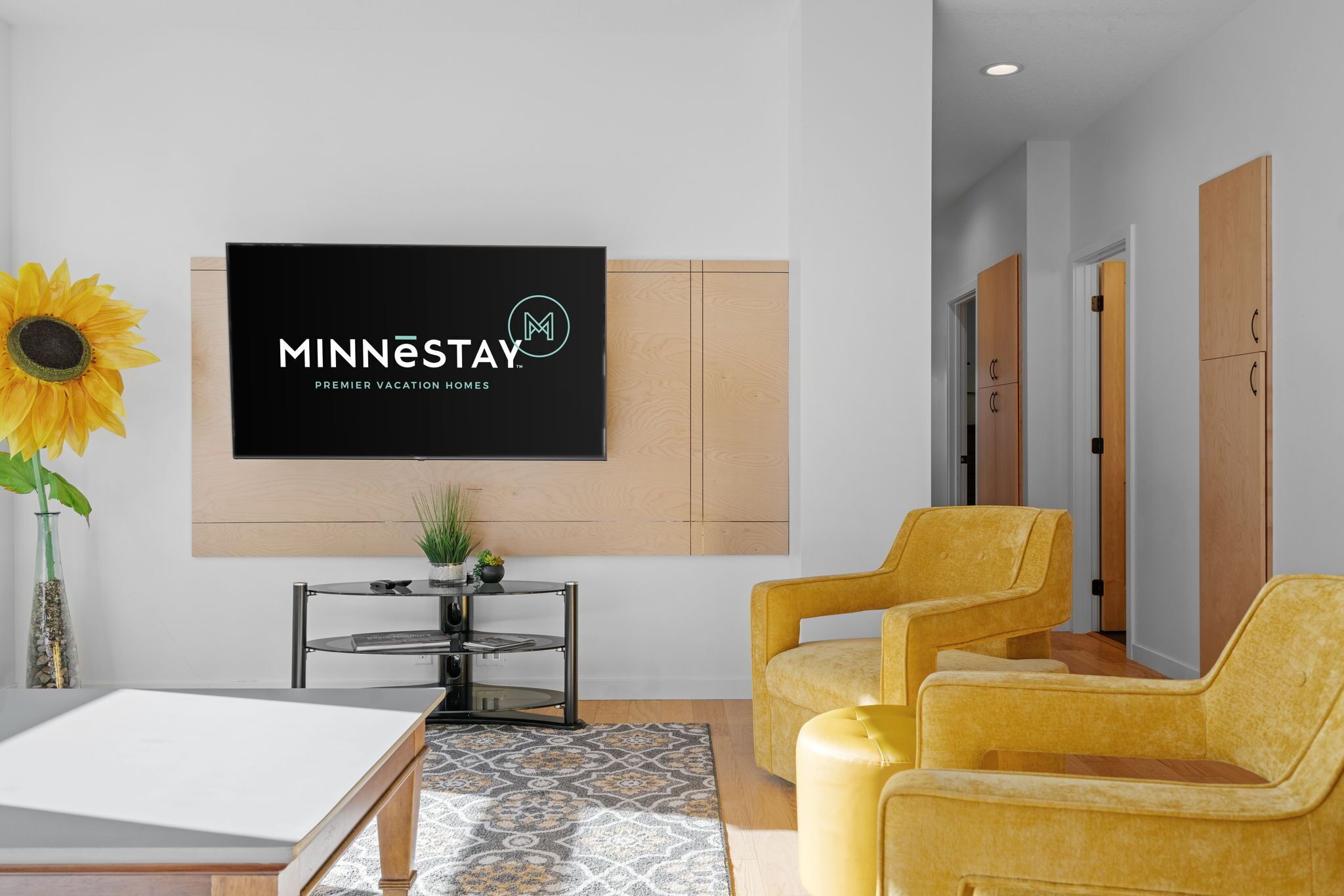 A modern hotel lobby with a large screen displaying the brand name, a round table with a vase and plant, and two yellow armchairs.