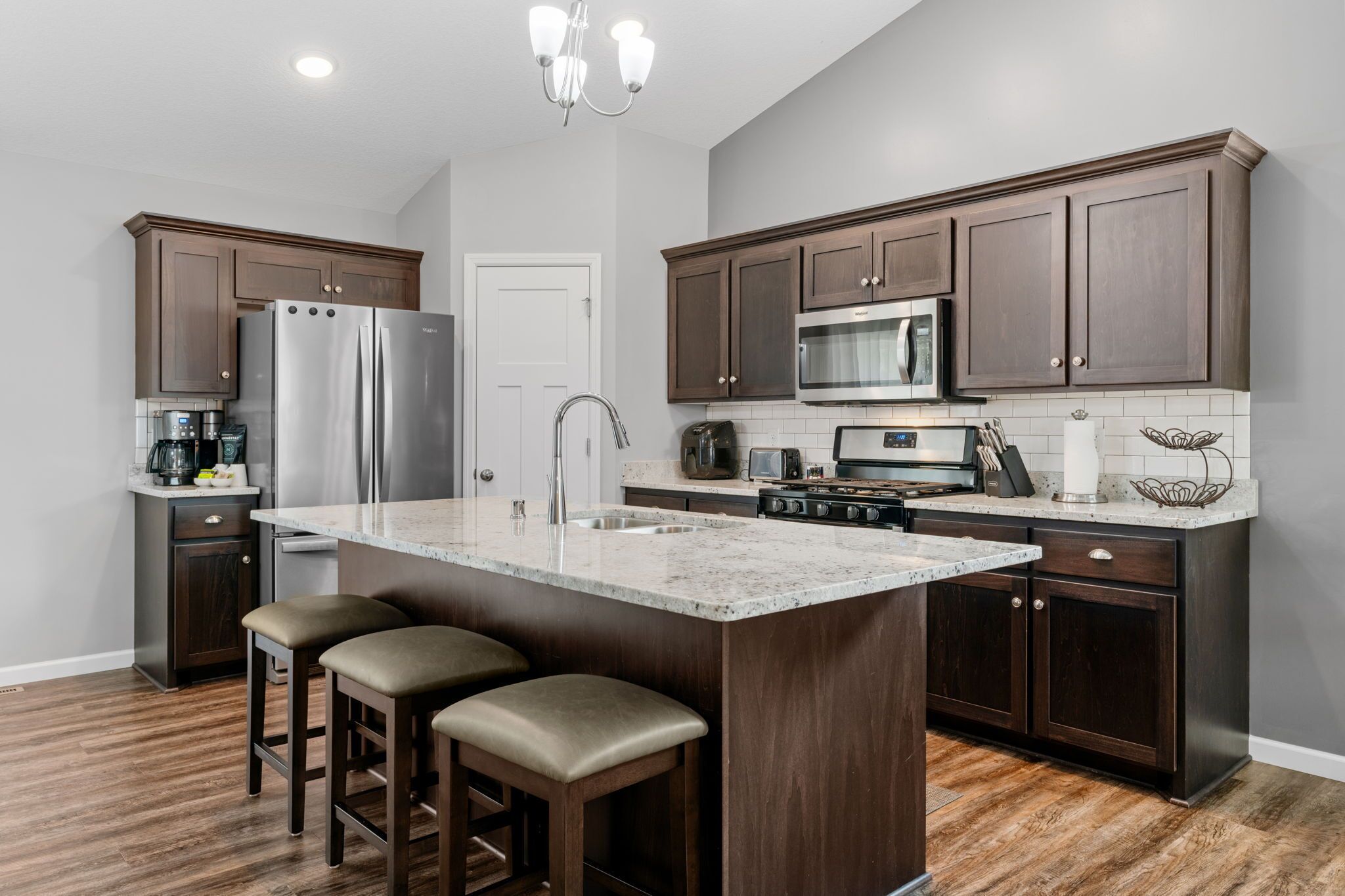 A modern kitchen with dark wood cabinets, stainless steel appliances, a large island with stools, and a tiled backsplash.