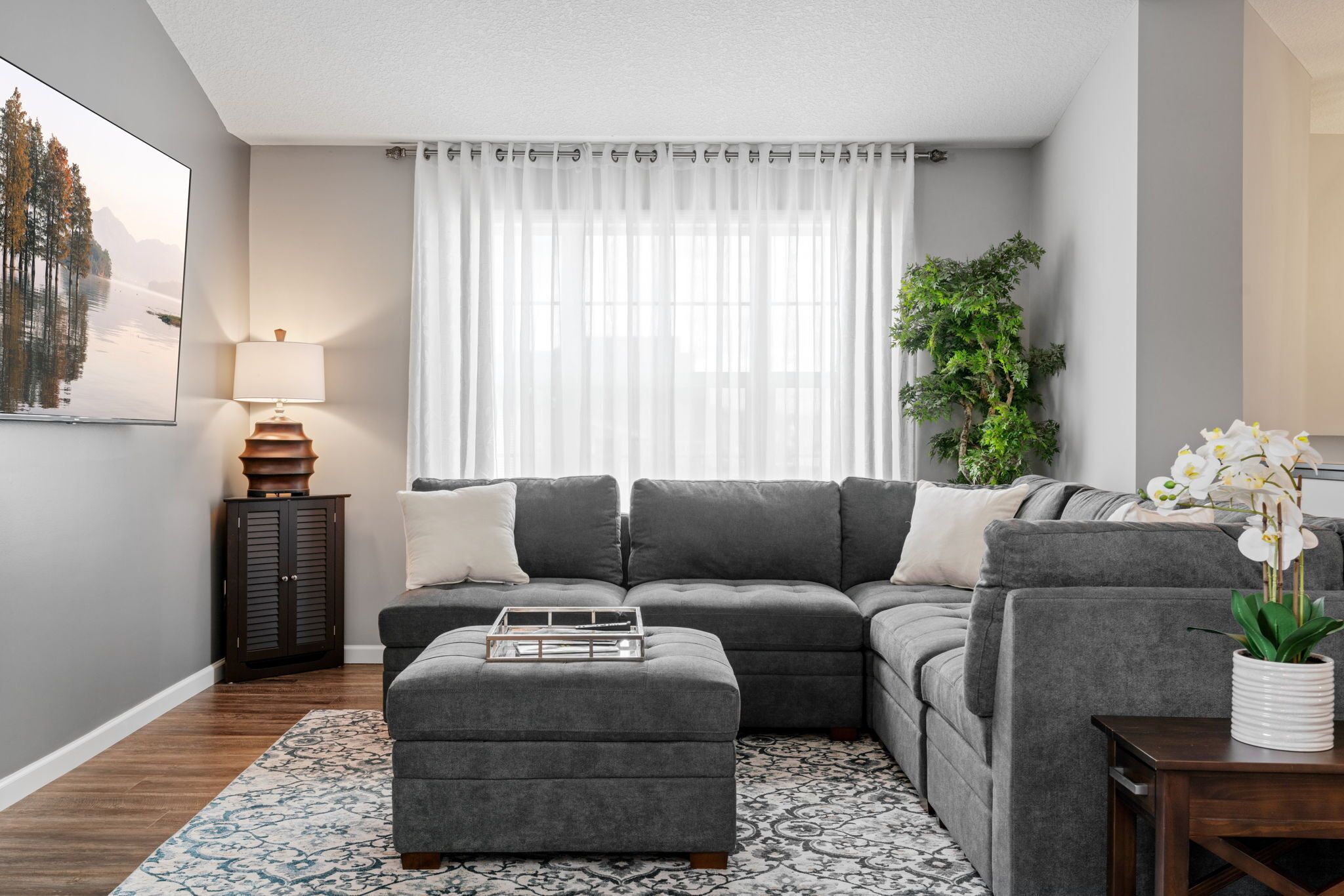 A living room with a sectional sofa, a small ottoman, a lamp, a plant, and a framed picture on the wall.