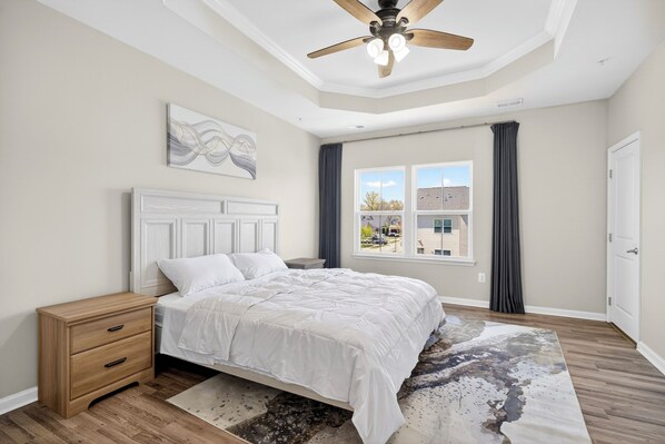 A bedroom with a bed, a nightstand, a ceiling fan, a window with curtains, and a view of buildings outside.