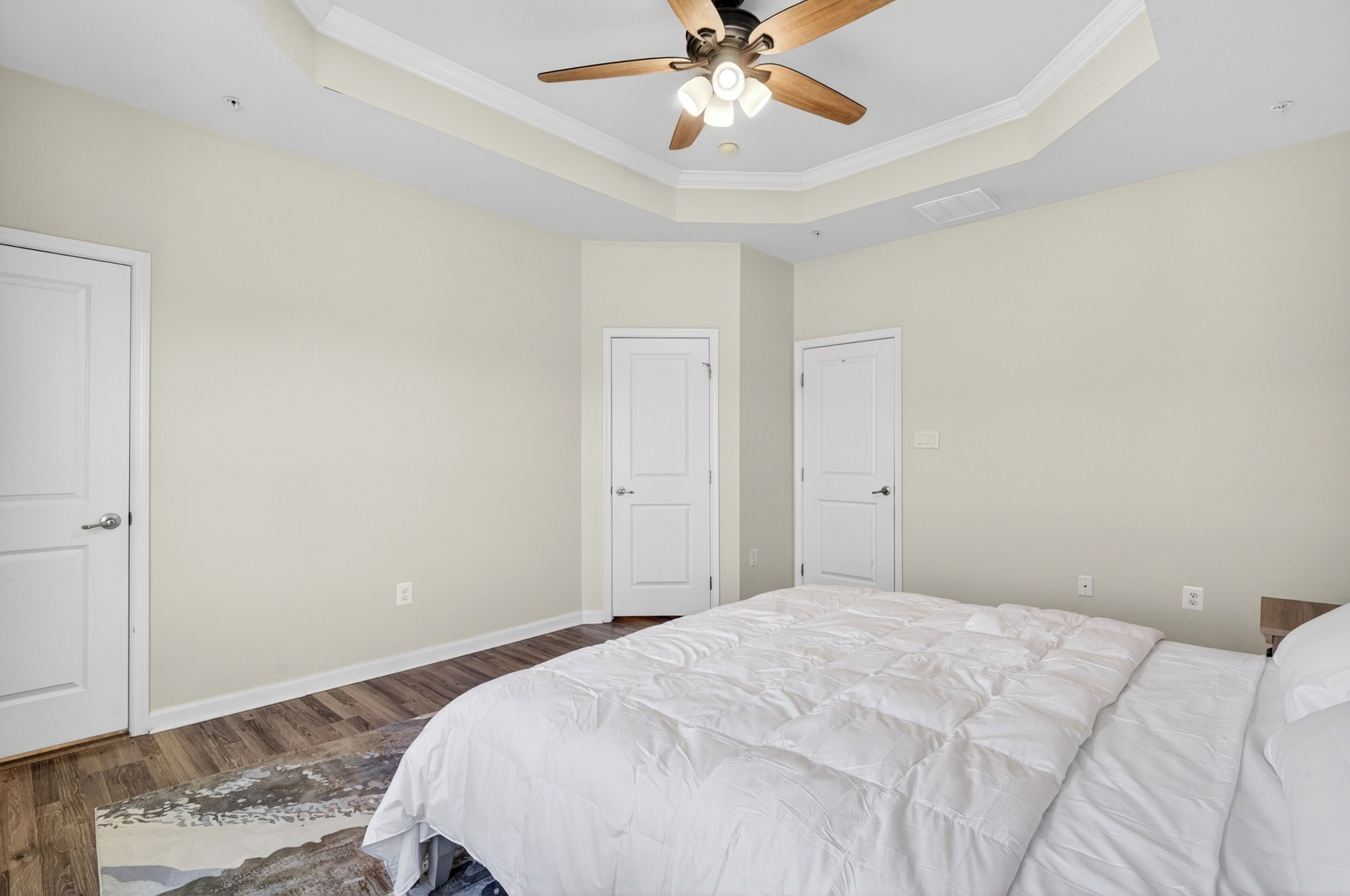 A bedroom with a large bed, two white doors, a ceiling fan, and a wooden floor.