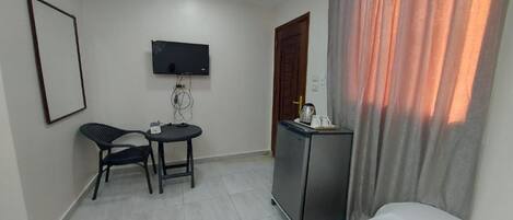 A hotel room with a bed, a desk with a chair, a TV mounted on the wall, a refrigerator, and a door with a curtain.