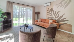 A living room with a sofa, two armchairs, a coffee table, and a TV on a stand. There is a large window with curtains, a floor lamp, and a wall with a leaf pattern. - Nice home in Oosterwolde with WiFi (Oosterwolde)