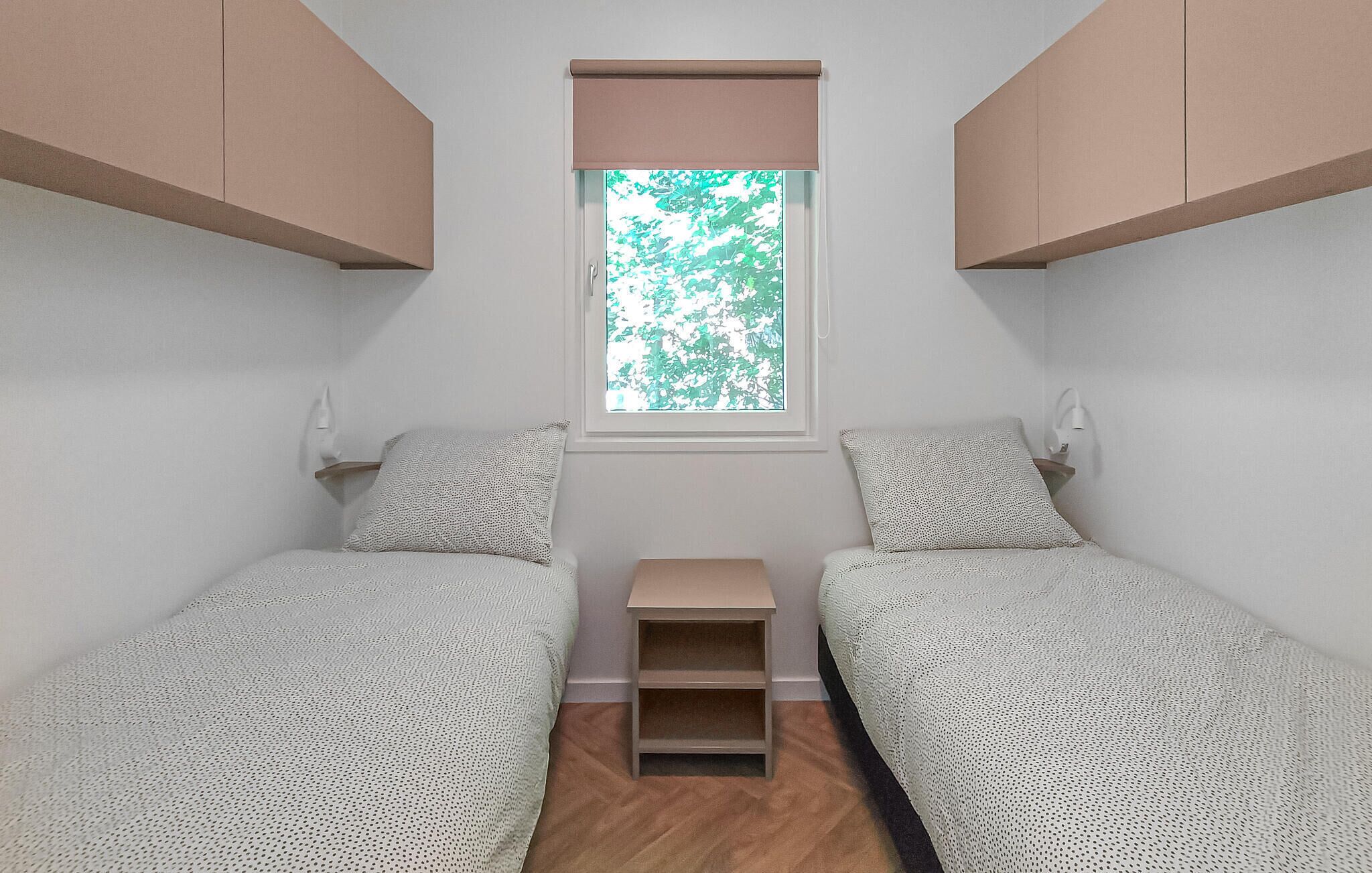Una habitación pequeña y minimalista con dos camas individuales, una ventana con vista a árboles y una pequeña estantería.