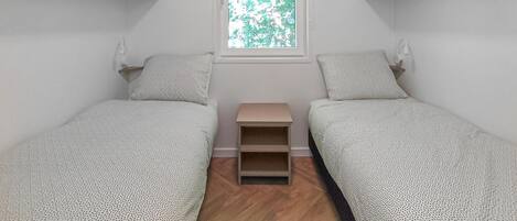 Una habitación pequeña y minimalista con dos camas individuales, una ventana con vista a árboles y una pequeña estantería.