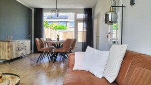 Una sala moderna con un sofá de cuero, una mesa de comedor de madera con sillas, una zona de cocina con un armario de madera y un ventanal con vistas a un patio trasero.