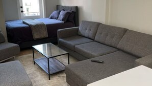 A modern living room with a sofa, a smaller armchair, a glass-top coffee table, and a bed with a dark-colored duvet.