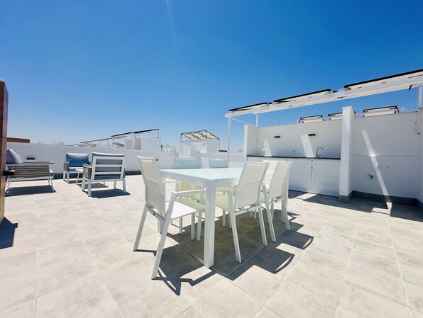 Une terrasse sur le toit avec une table et des chaises blanches, un bar intégré et un ciel d’un bleu limpide.