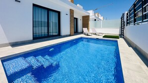 Une piscine extérieure moderne, avec une eau d’un bleu limpide, entourée d’un bâtiment blanc doté de grandes portes-fenêtres coulissantes et d’un mur d’accent en bois.