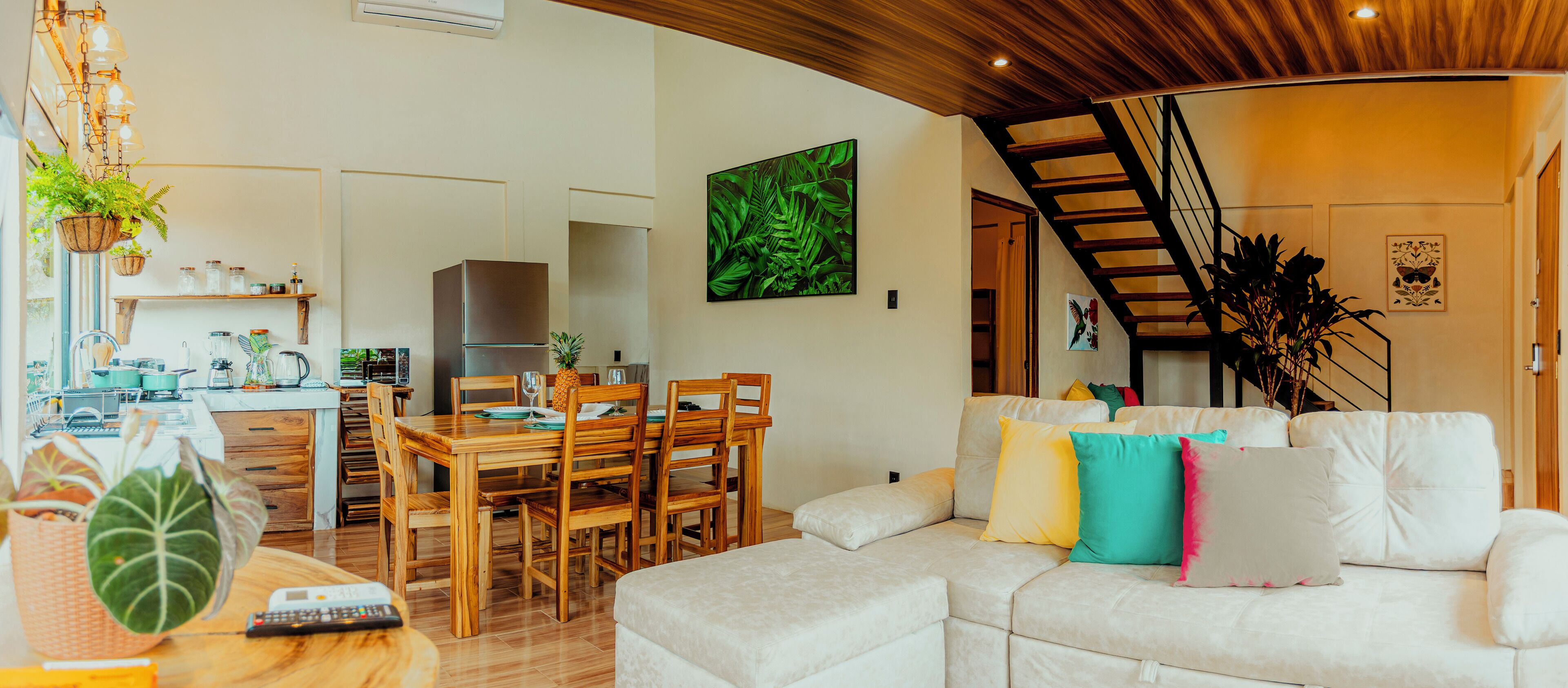 Uma sala de estar moderna com sofá branco, mesa de jantar de madeira e cadeiras. Há uma escada que leva a outro andar, e uma área de cozinha com geladeira e uma planta.