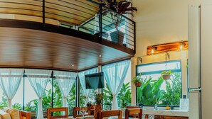 A spacious dining area with a wooden table and chairs, a television mounted on the wall, and large windows with curtains. - Cabañas Caeli, El Colibri, NEW La Fortuna Rainforest Cabin w Panoramic Views (Peñas Blancas)