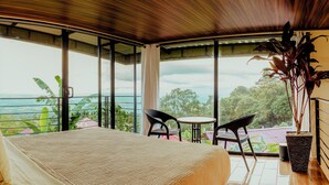 A bedroom with a large window, a bed, a nightstand, and a potted plant. - Cabañas Caeli, El Colibri, NEW La Fortuna Rainforest Cabin w Panoramic Views (Peñas Blancas)