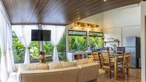 A spacious living area with a sofa, wooden dining table, and chairs. There is a kitchen area with a refrigerator, microwave, and sink. Large windows provide views of greenery outside. - Cabañas Caeli, El Colibri, NEW La Fortuna Rainforest Cabin w Panoramic Views (Peñas Blancas)
